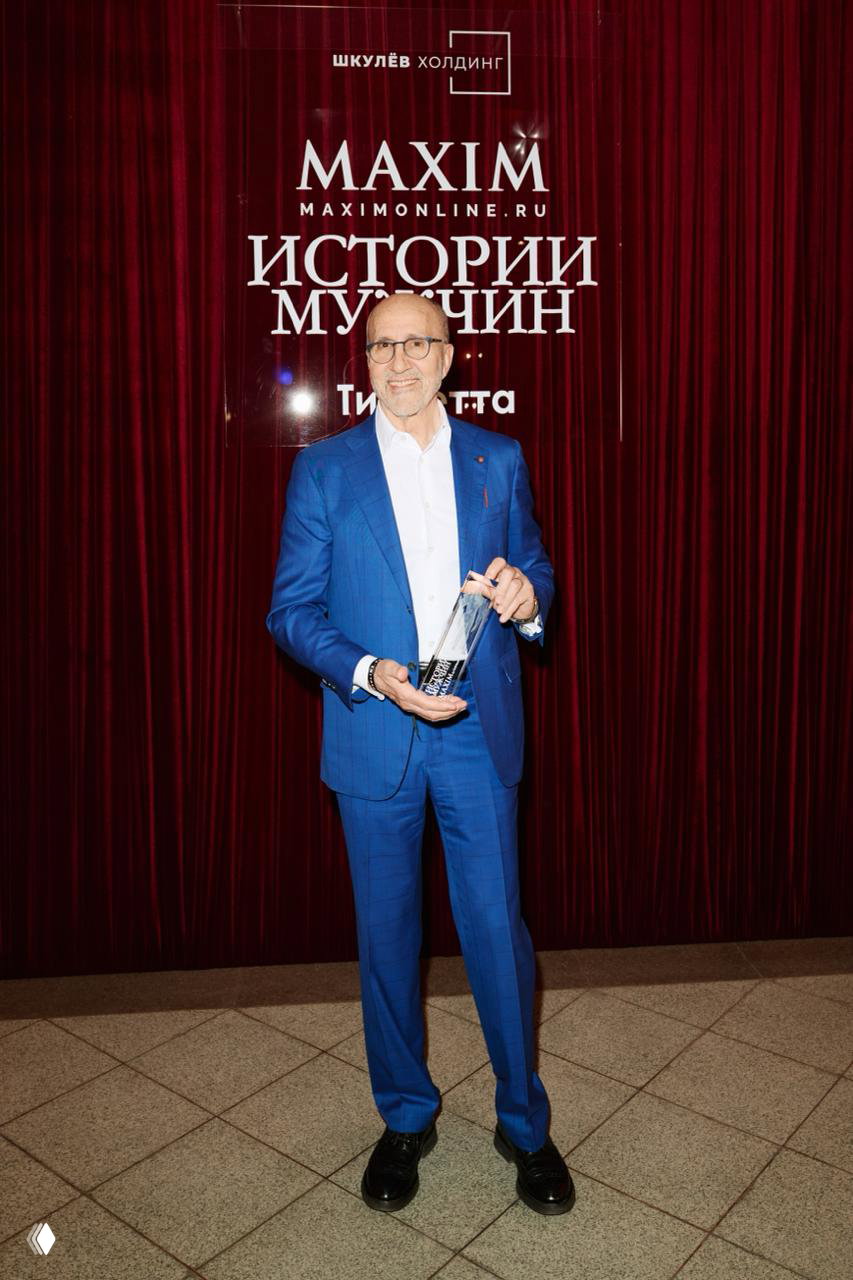 Ещё один гость вечера в синем костюме позирует с хрустальной статуэткой на фоне логотипа Maxim и декоративной шторы.