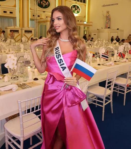 Участница от России в розовом платье с лентой «RUSSIA» и небольшим флажком позирует в банкетном зале на фоне сервированных столов.