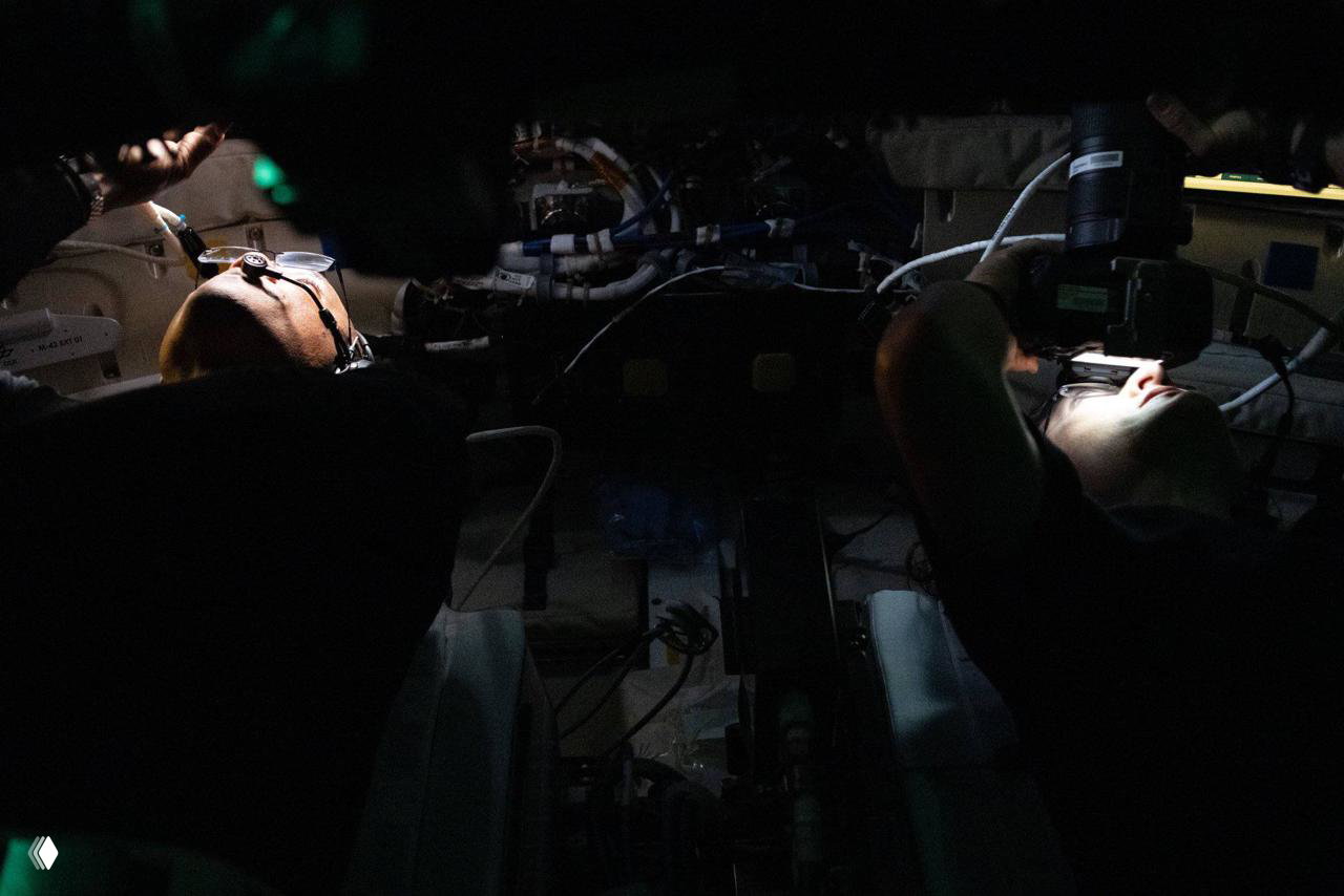 Астронавты работают в затемнённом отсеке, локальная подсветка подчёркивает оборудование и инструменты; документальный кадр рабочей рутины во время миссии.