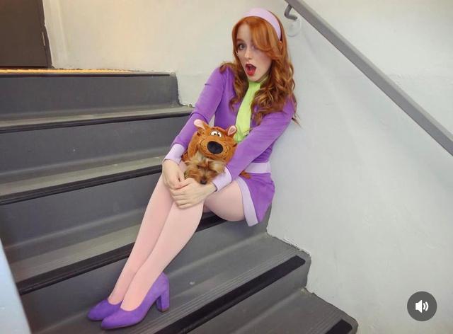 Актриса в костюме Дафны сидит на ступеньках и держит мягкую игрушку Скуби‑Ду — кадр со съёмочной репетиции или фан‑косплея, заметна фиолетовая одежда.