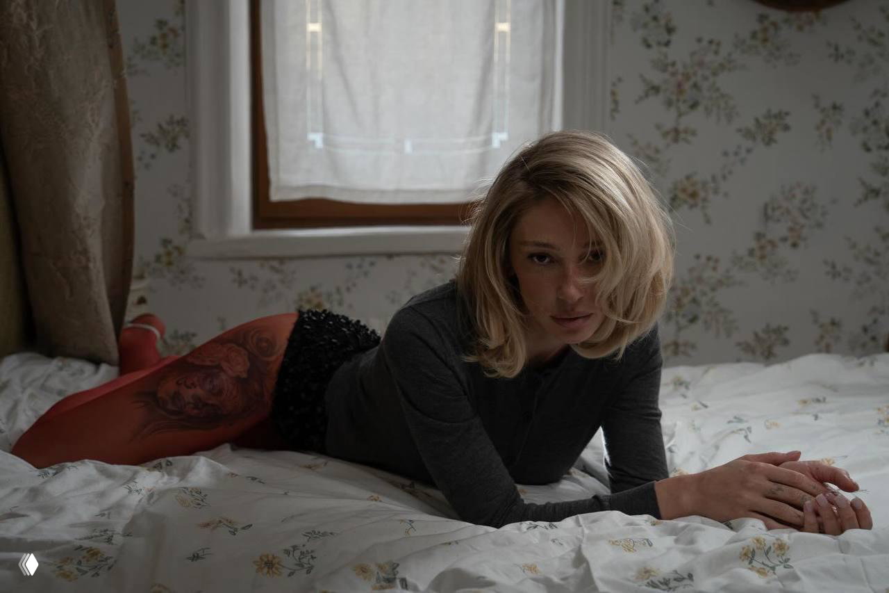 Женщина лежит на животе на кровати в тонких красных колготках, на бедре заметна крупная татуировка; кадр в естественном освещении.