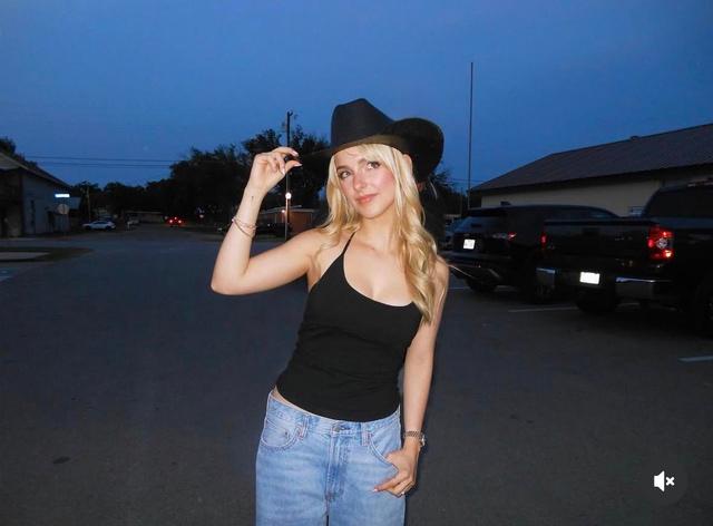 Портрет блондинки в джинсах и чёрной майке на улице вечером — возможно фотография актрисы вне съёмочной площадки, фон парковка и машины.