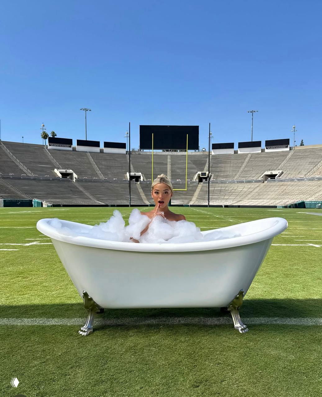 Модель в наполненной пеной белой ванне на газоне стадиона, на фоне ворот и широких пустых зрительских трибун под чистым небом.