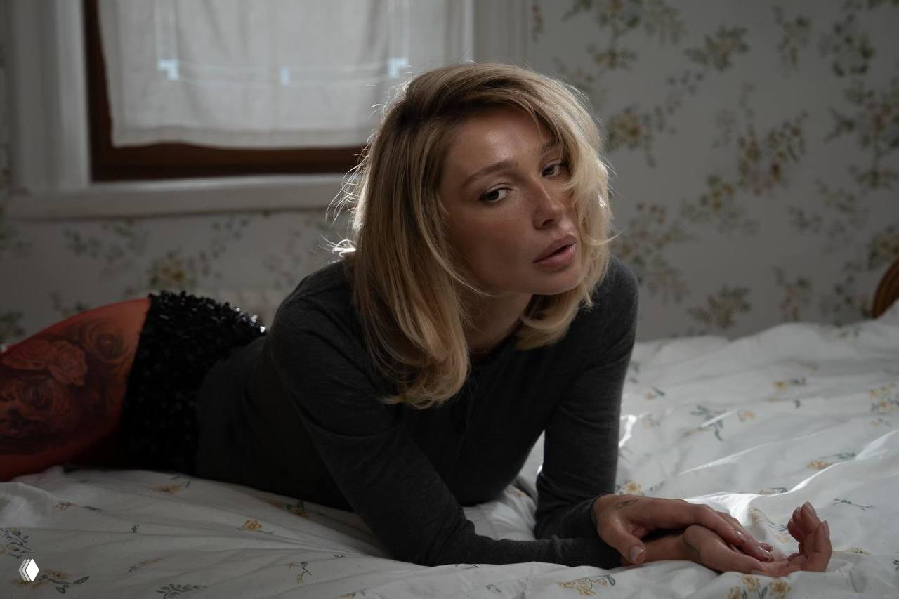 Полублизкий портрет женщины на кровати: светлые волосы, тёмный свитер и красные колготки на фоне винтажного интерьера и окна.