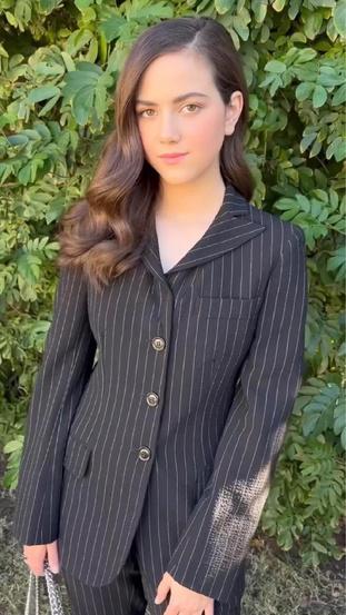 Портрет молодой темноволосой актрисы в пиджаке на фоне зелёной листвы — вероятный каст‑фото или пресс‑снимок, спокойный нейтральный фон.