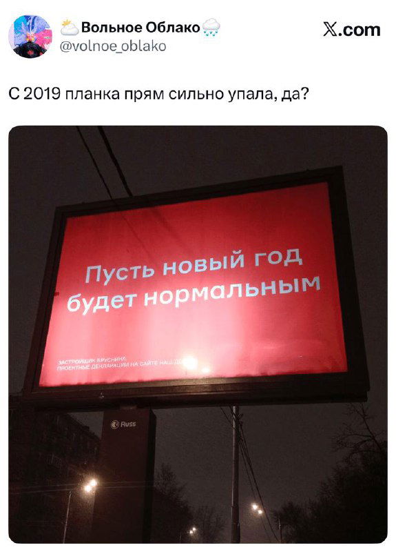 Фотография уличного билборда с красным фоном и белой надписью «Пусть новый год будет нормальным», ночной городской кадр с рекламой и уличным освещением вокруг, ироническая вывеска.