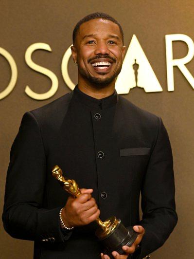 Улыбающийся мужчина в тёмном костюме держит статуэтку «Оскар» на фоне эмблемы церемонии — праздничный портрет победителя.