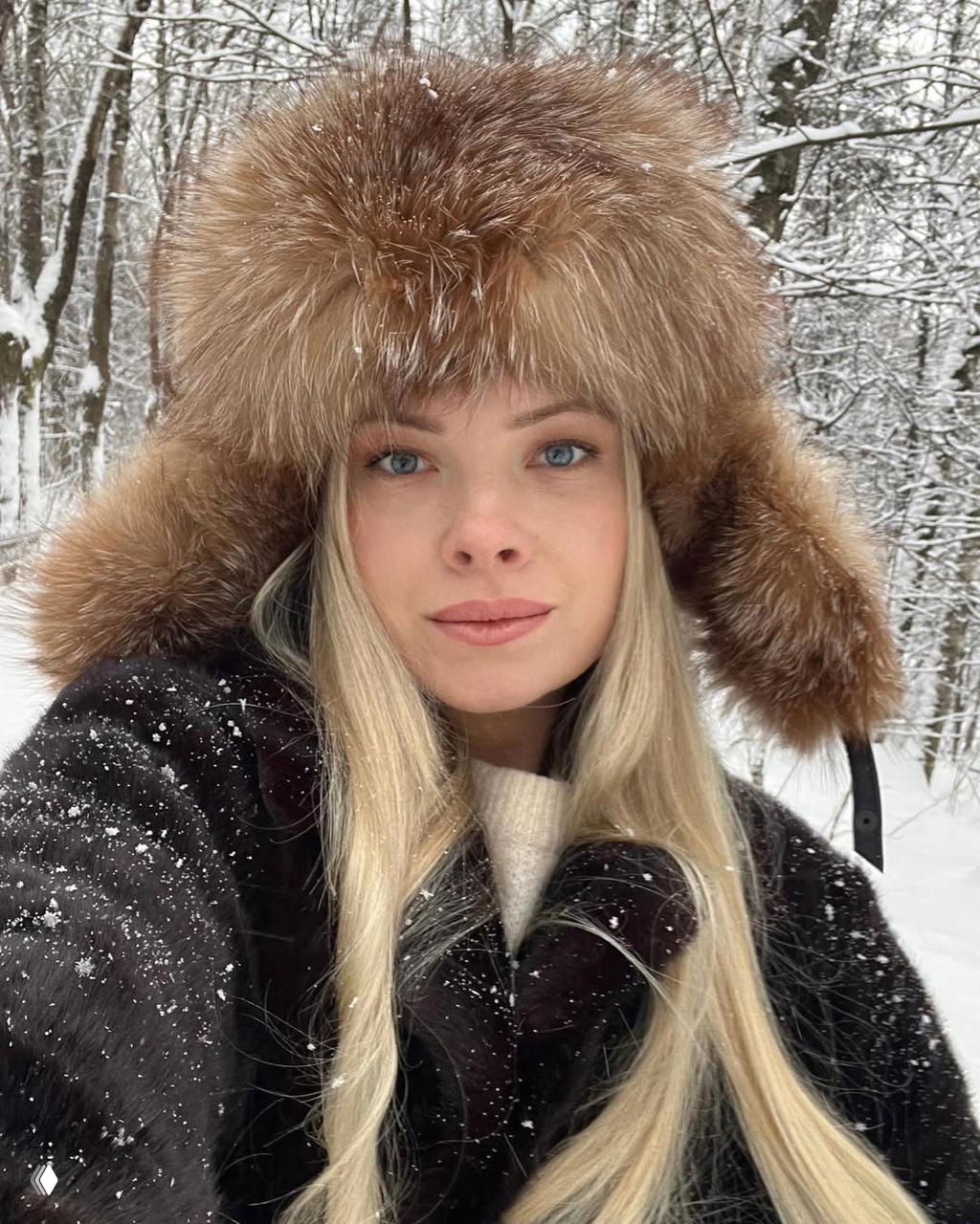 Женский портрет на фоне зимнего леса: блондинка в тёплой шапке из меха и тёмной шубе, снег на волосах и одежде, суровый северный образ.