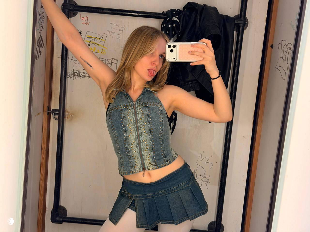 Молодая женщина делает селфи в примерочной или гримерной: на ней джинсовый корсет и мини-юбка, поза перед зеркалом, в углу видна вешалка с одеждой.