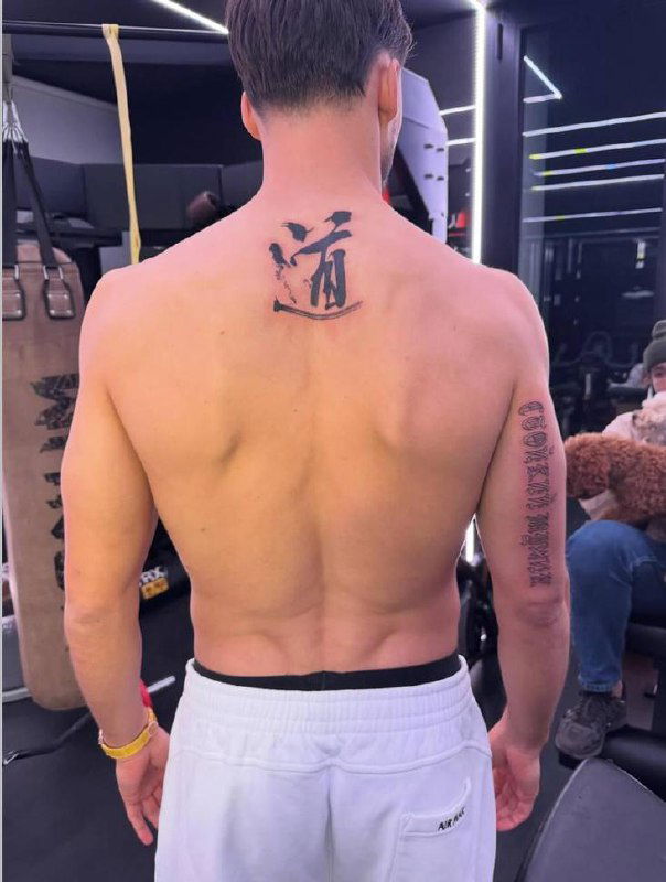 Татуировка на верхней части спины футболиста: чёрные готические буквы с фразой «стойкий мужик», снимок сделан в тату‑студии, вид сзади.
