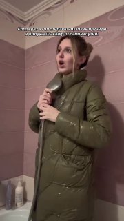 Девушка в зелёном пуховике стоит в комнате с розовыми обоями и эмоционально держит куртку у горла — кадр из видео о реакции на фетиш.