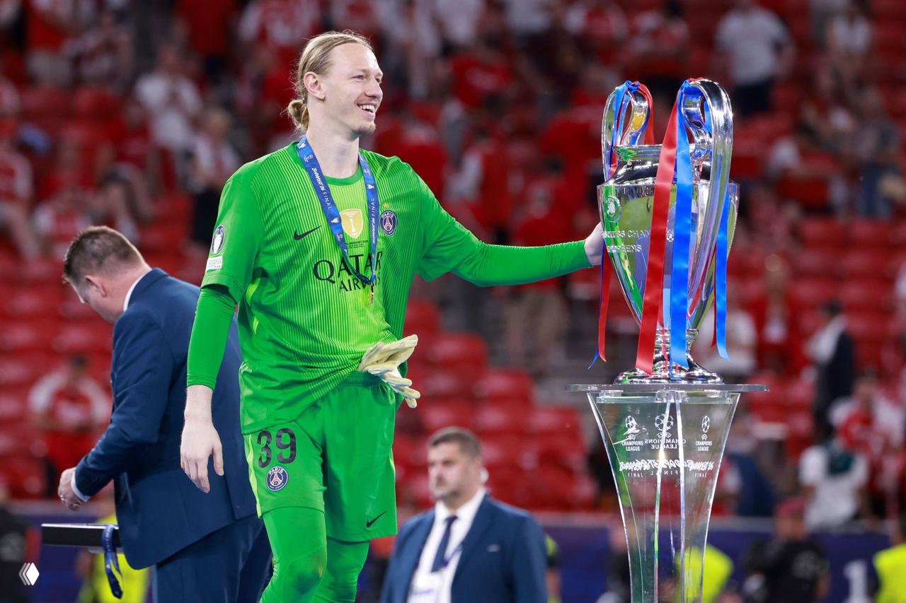Матвей Сафонов в зелёной игровой форме держит большой трофей Лиги чемпионов на поле, вокруг видны официальные лица и трибуны.