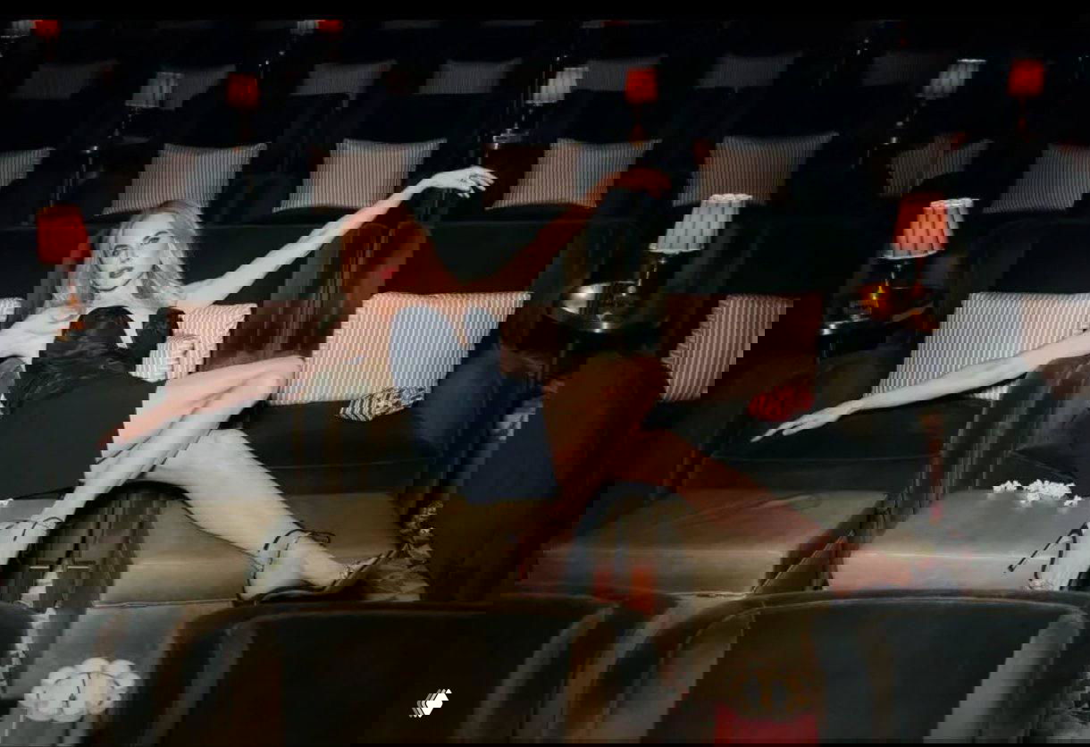 Женская фигура в кинозальном интерьере: чёрное вечернее платье, туфли на каблуке, расслабленная поза на бархатных креслах, бокалы и попкорн в кадре — стильная съёмка.