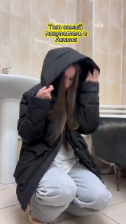 Молодая женщина в чёрном пуховике присела в помещении, держит капюшон, видны светлые штаны и интерьер, снимок выглядит как короткое видео в соцсетях.