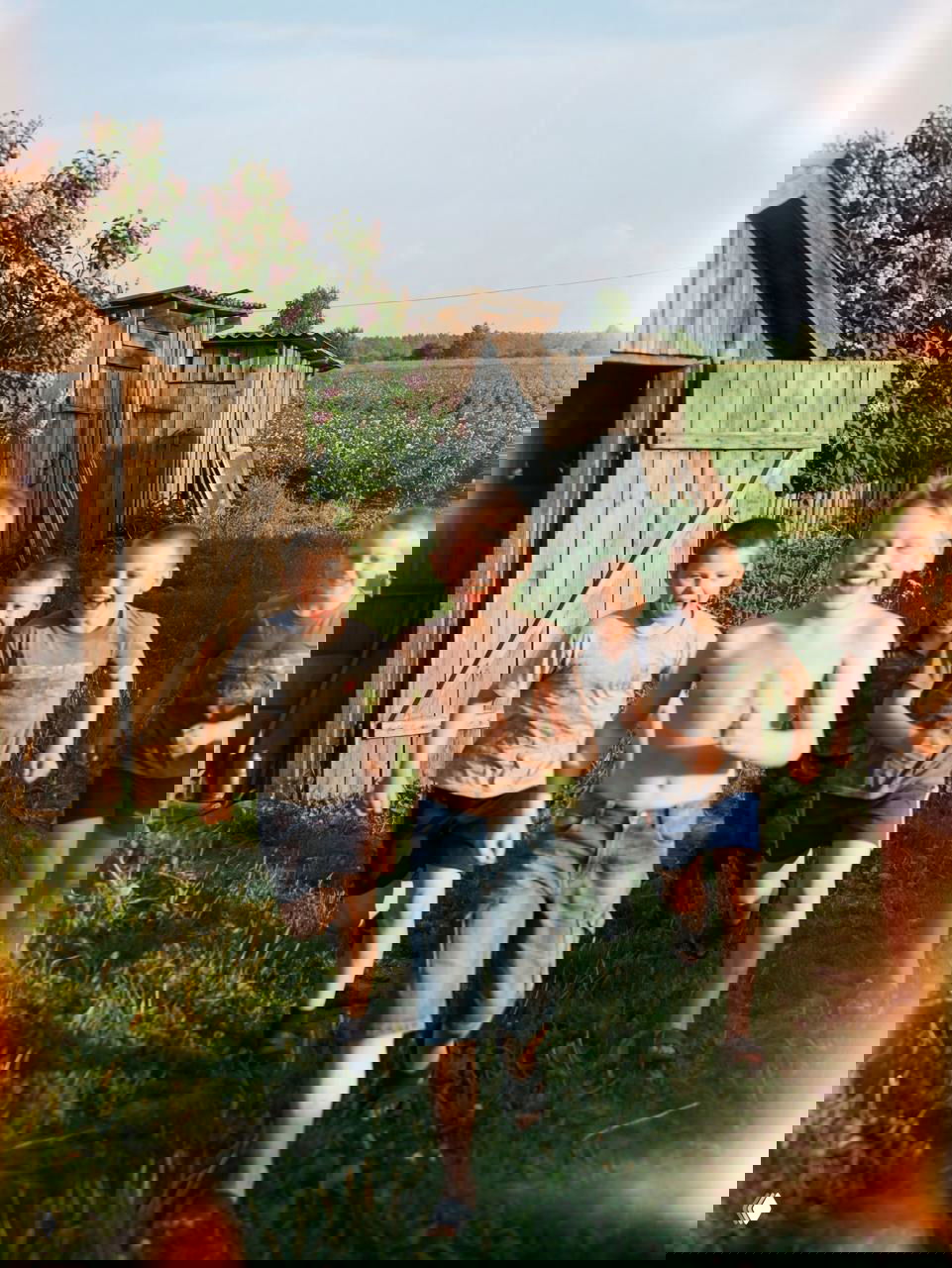 Группа ребят бежит по двору на фоне сельских туалетов, сараев и широкого поля, летнее солнце и искрящиеся лучи — живые эмоции детства.