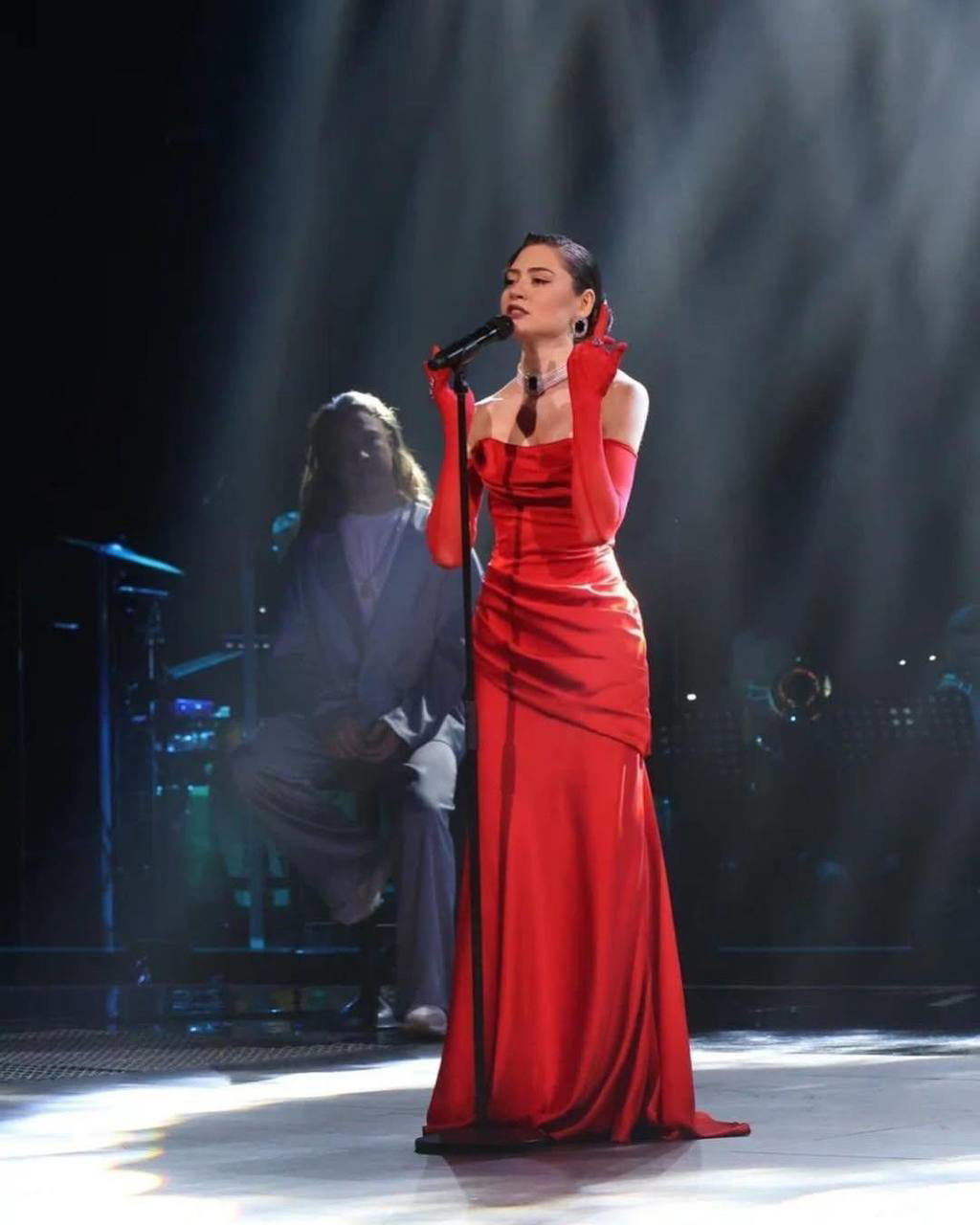 Исполнительница в длинном красном платье выступает на сцене с микрофоном, театральное освещение и оркестр позади