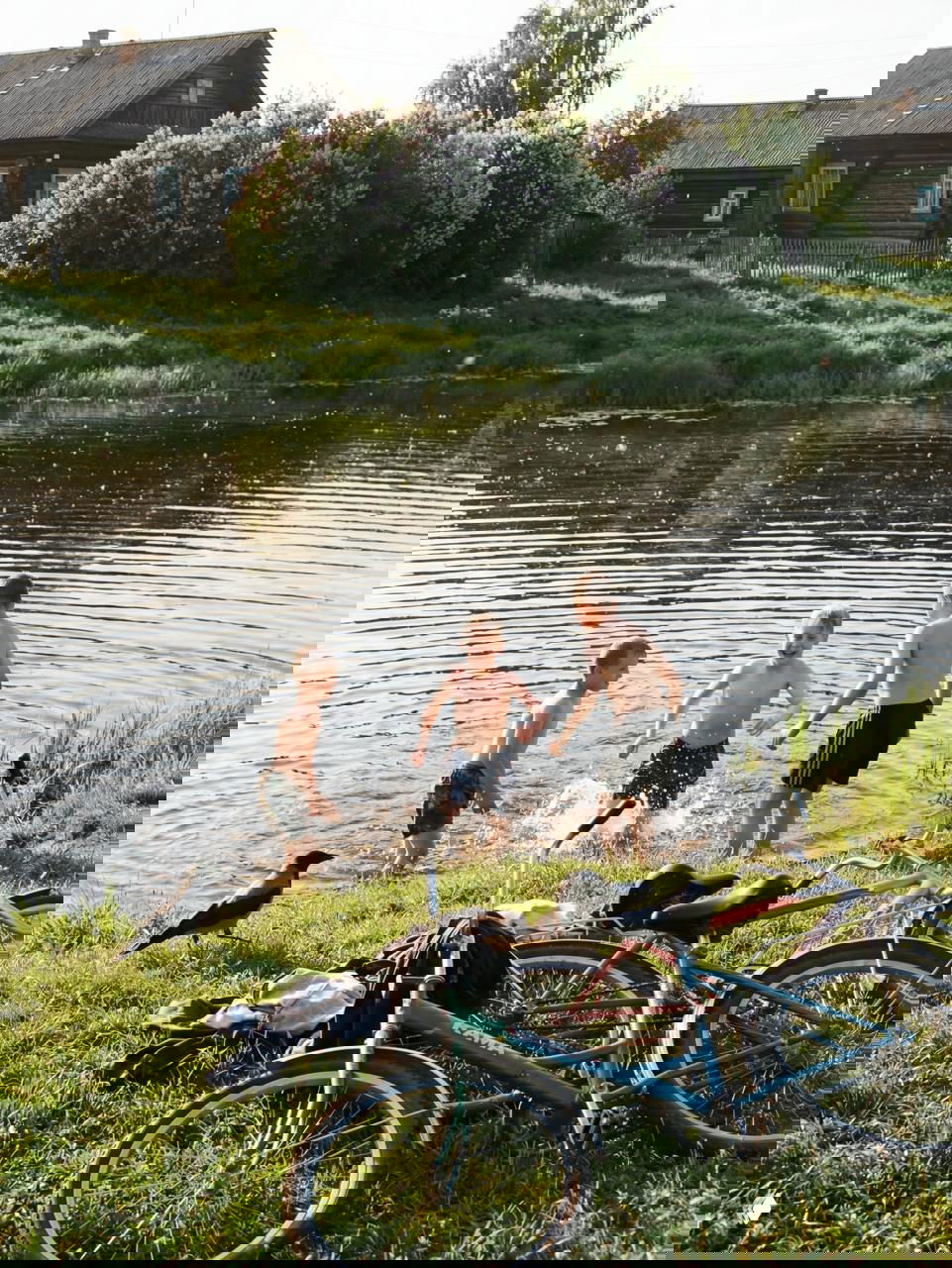 Трое мальчиков плескаются в тихом деревенском пруду; на переднем плане несколько старых велосипедов, прибрежная трава и летние отражения воды.