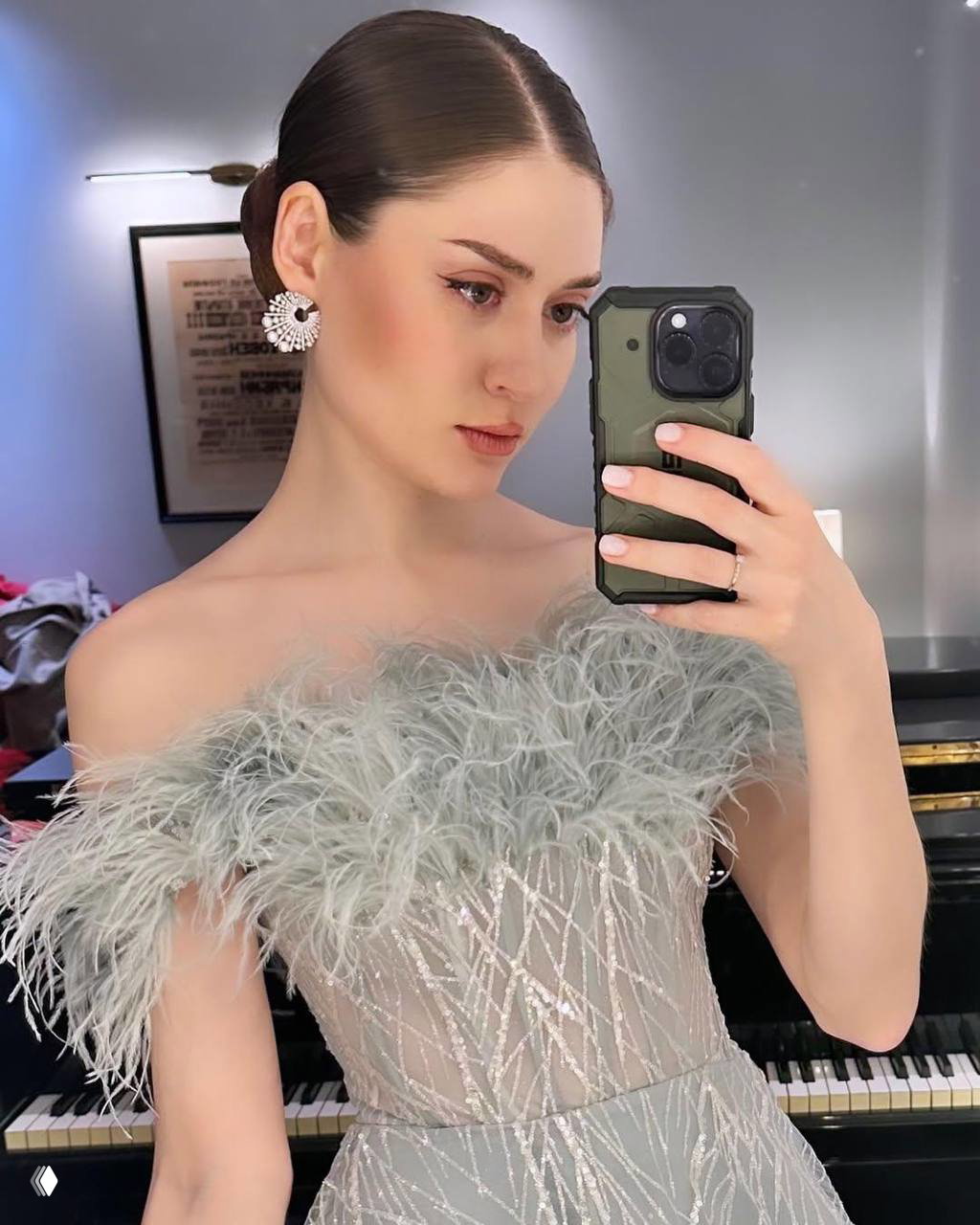 Крупный план в гримерке: девушка делает селфи у рояля в платье с перьями, видно телефон в руке и серьги крупного плана