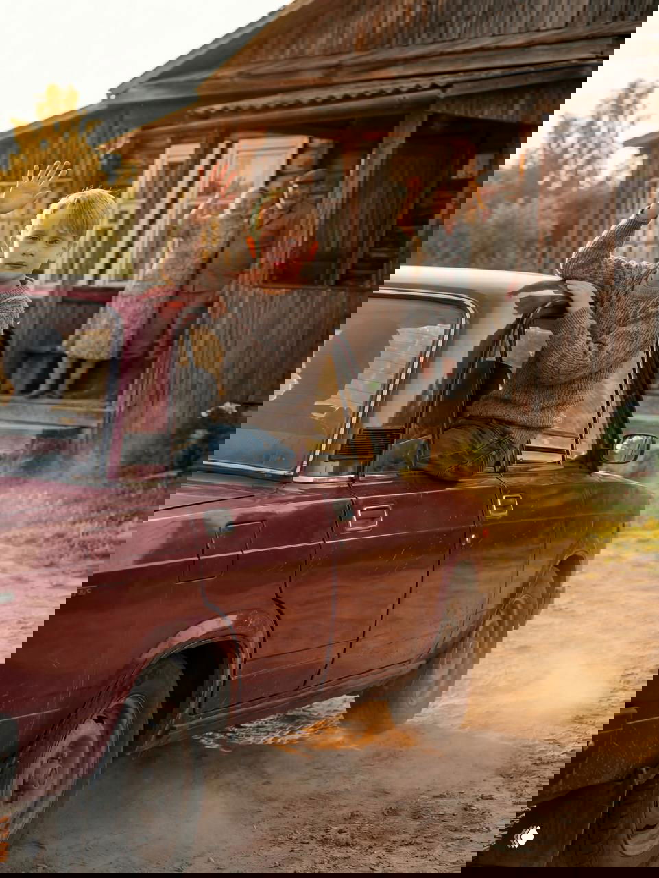 Мальчик сидит на двери старой машины и машет рукой, позади на крыльце пожилая пара прощается; пыльная деревенская дорога на закате создаёт ностальгическое настроение.