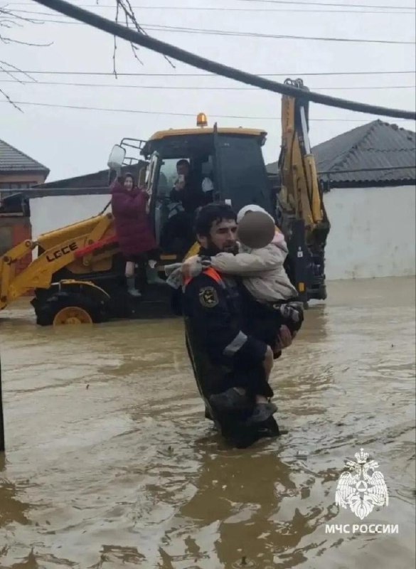 Спасатель несёт ребёнка на руках через воду, за ним экскаватор помогает в эвакуации; уличная сцена затопления и люди на технике.