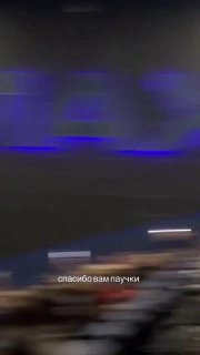Вертикальное видео из зала кинотеатра: тёмный экран с синей подсветкой, размытые контуры зрителей внизу и световые блики.