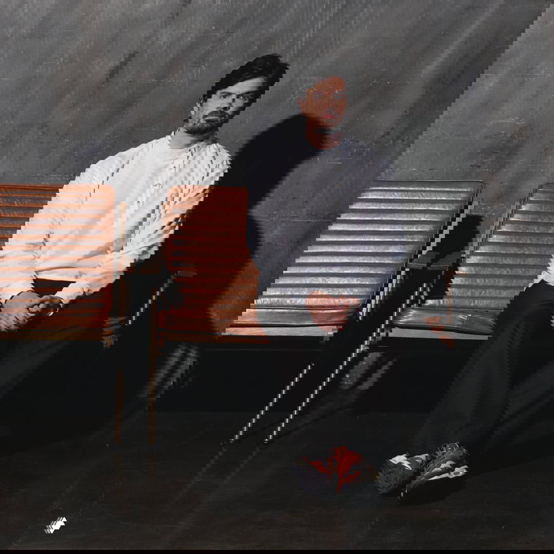 Мужчина в белой косоворотке сидит на кожаном кресле в студийном интерьере; образ подчёркивает рубашку из коллаборации бренда.