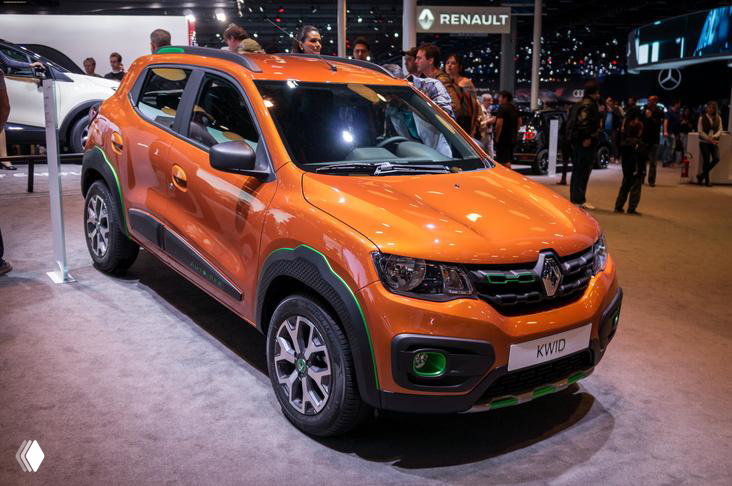 Оранжевый Renault Kwid на автомобильной выставке, снят под углом, машина на экспозиции с посетителями и стендами на заднем плане.