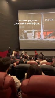 Широкий кадр зала с группой гостей в масках и костюмах спайдермена у экрана, часть зрителей в креслах снимает происходящее на телефон.