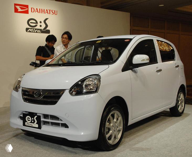 Белый компактный Daihatsu Mira e:S на презентации, демонстрационный стенд с логотипом марки и людьми рядом, вид вблизи.