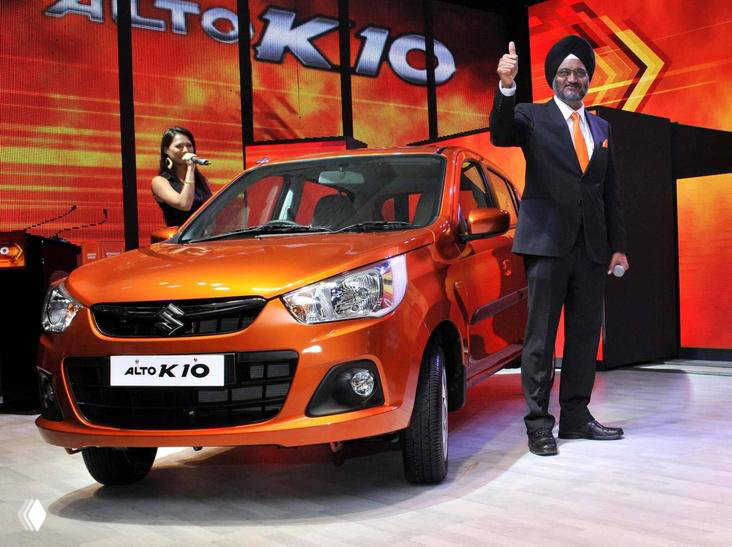 Оранжевый Maruti Suzuki Alto на сцене презентации, рядом ведущий и декорации, машина показана в торжественной обстановке.