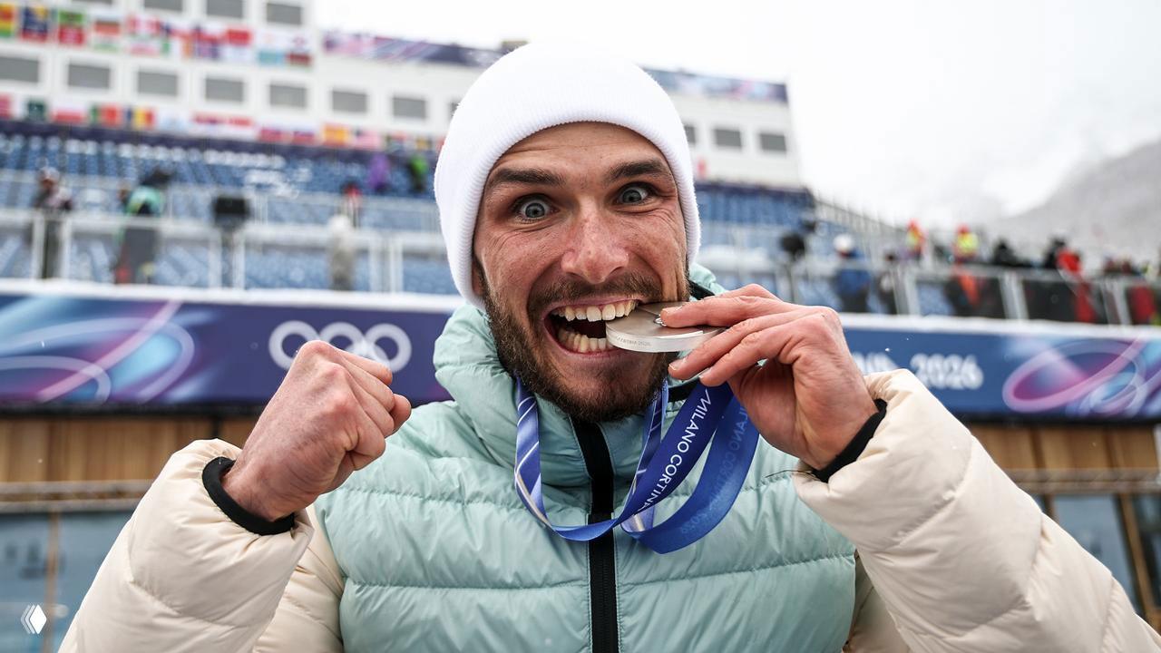 Никита Филиппов кусает серебряную медаль на фоне трибун Олимпиады‑2026, радостное выражение лица и жест победы