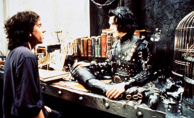 Кадр из сцены в интерьере: персонаж в мрачном костюме сидит за столом в мастерской, вокруг книги и механизмы декора.