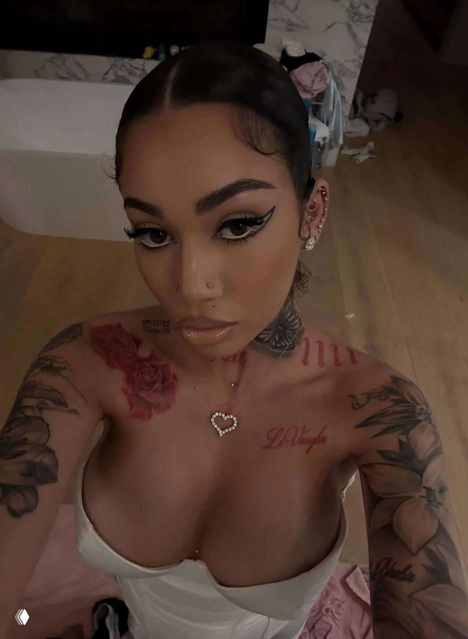 Селфи модели с тёмными причёской и татуировками, кадр в домашней обстановке подчёркивает стиль и детали тела модели.