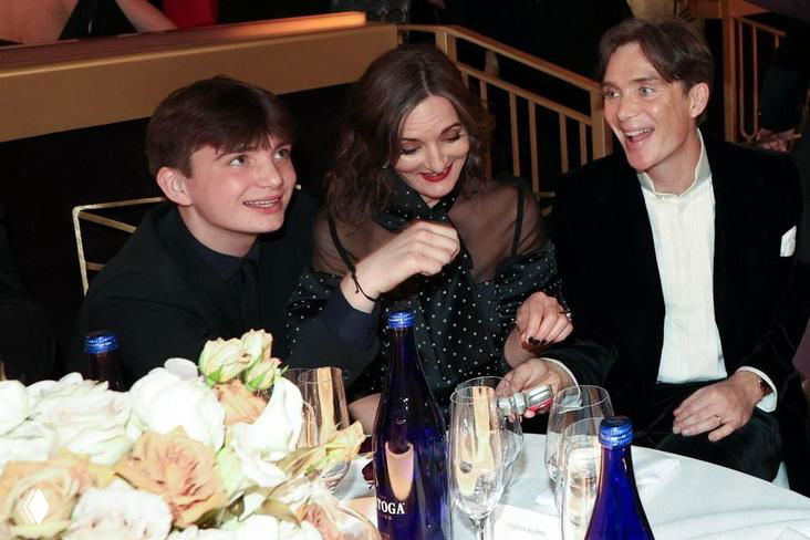 Сцена за столом: женщина, мужчина и молодой парень сидят рядом за праздничным столом, на столе цветы, бутылки и бокалы, тёплая обстановка.