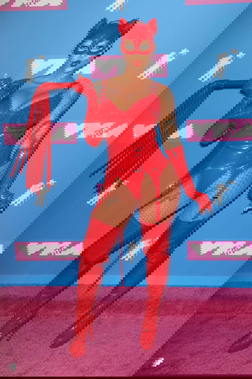 Профессиональное фото в латексном костюме красного цвета на фоне брендовой фотозоны, образ сценический и провокационный, подчёркнуты аксессуары.