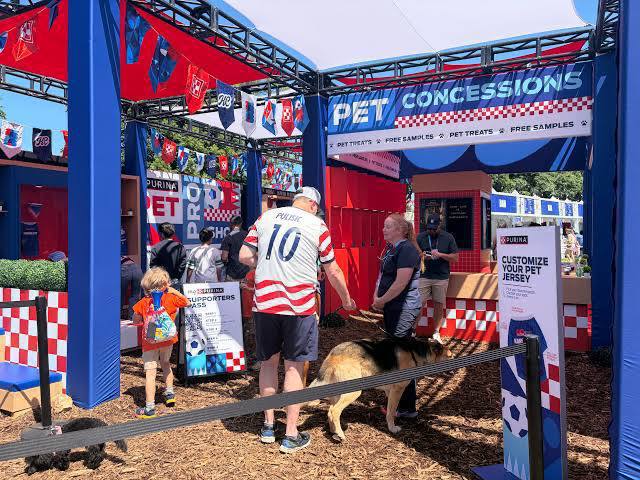 Вход в зону Pet Concessions на ЧМ-2026 в Канзасе: люди с собаками, флаги, киоски и указатели для владельцев питомцев в фан-зоне.