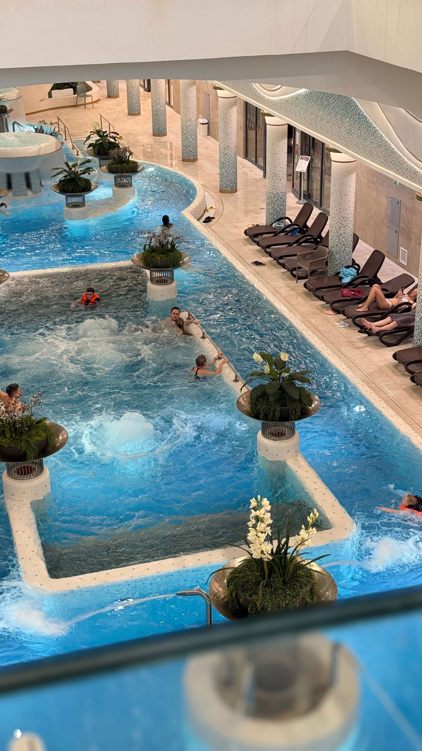 Вид сверху на внутренний термальный бассейн с насыщенно-голубой водой, джакузи и шезлонгами; люди отдыхают в современном спа-комплексе.