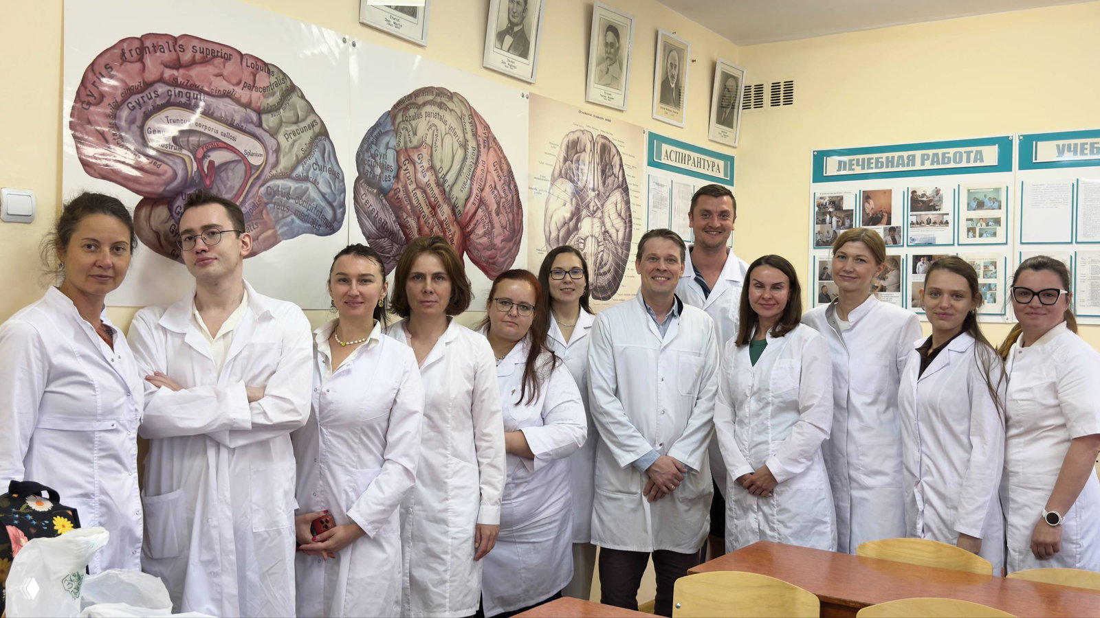 Группа медицинских сотрудников в белых халатах позирует в учебной аудитории на фоне больших постеров мозга и схем.