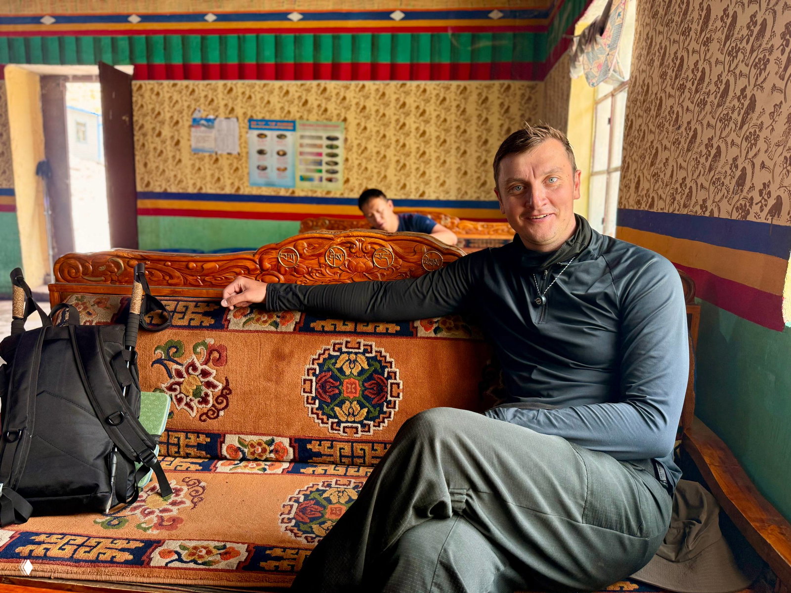 Внутреннее пространство тибетской гостиной: мужчина сидит на резном деревянном диване в комнате с цветными орнаментами и рюкзаком рядом.