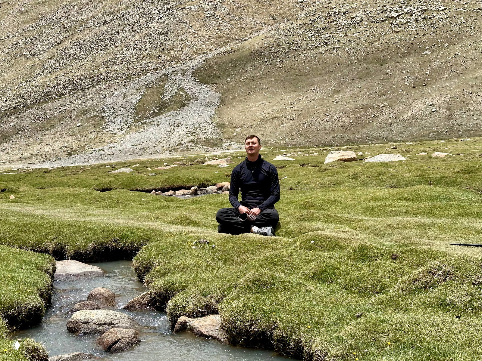 Мужчина медитирует, сидя на зелёной альпийской лужайке у небольшого ручья в высокогорье недалеко от Кайлаша, вокруг каменистые склоны.