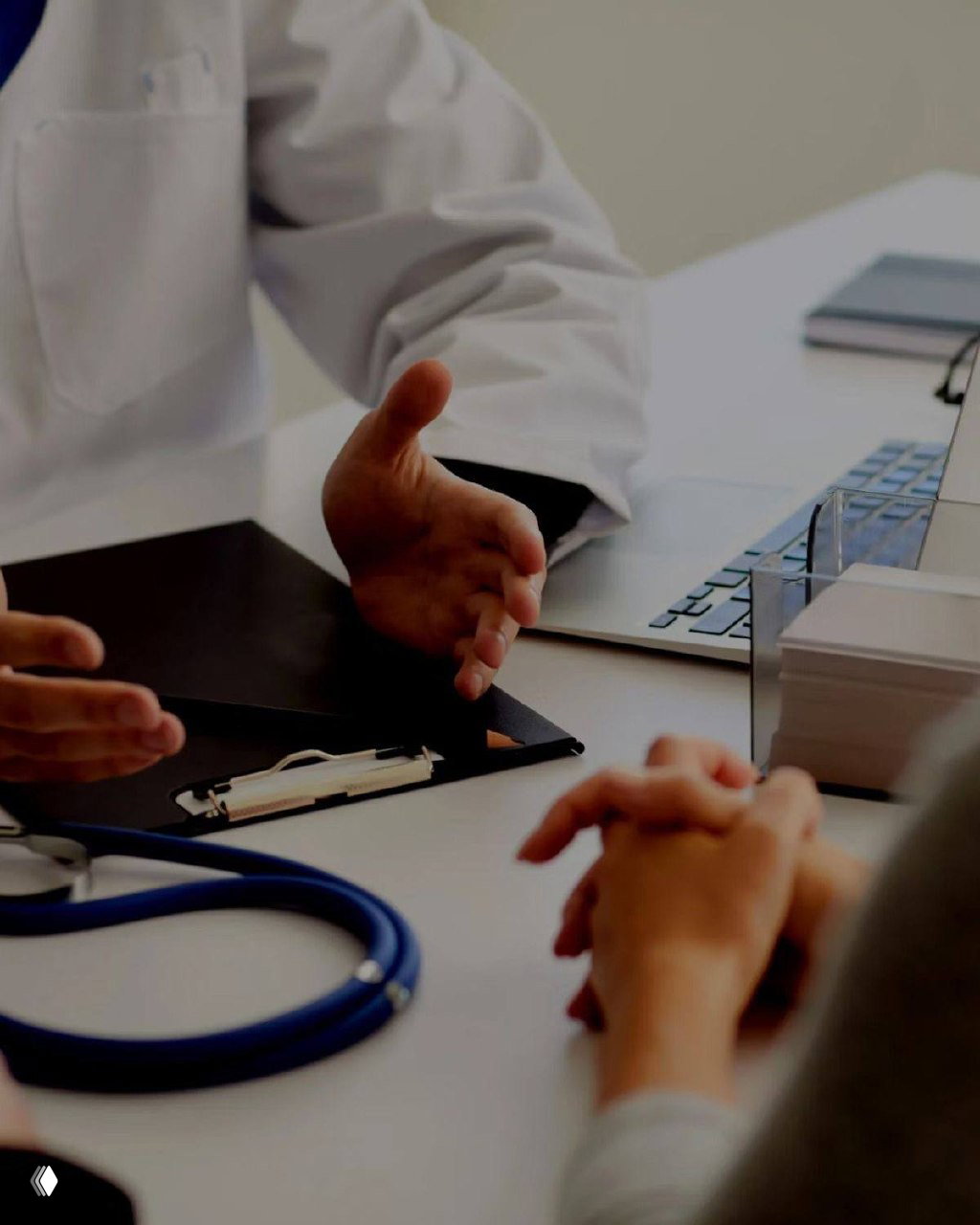 Врач в белом халате за столом объясняет результаты обследования пациенту; на столе стетоскоп, планшет и папка с документами, руки пациентов видны в кадре.