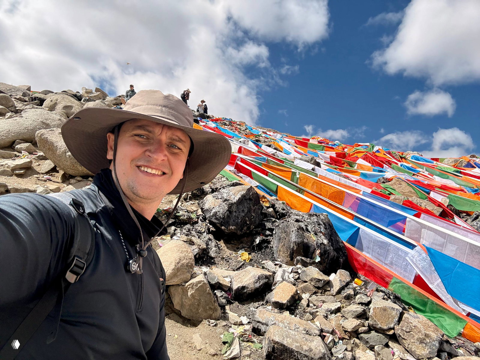 Селфи автора на фоне холмов, покрытых яркими тибетскими молитвенными флажками, небо с облаками и каменистый склон Кайлаша позади.