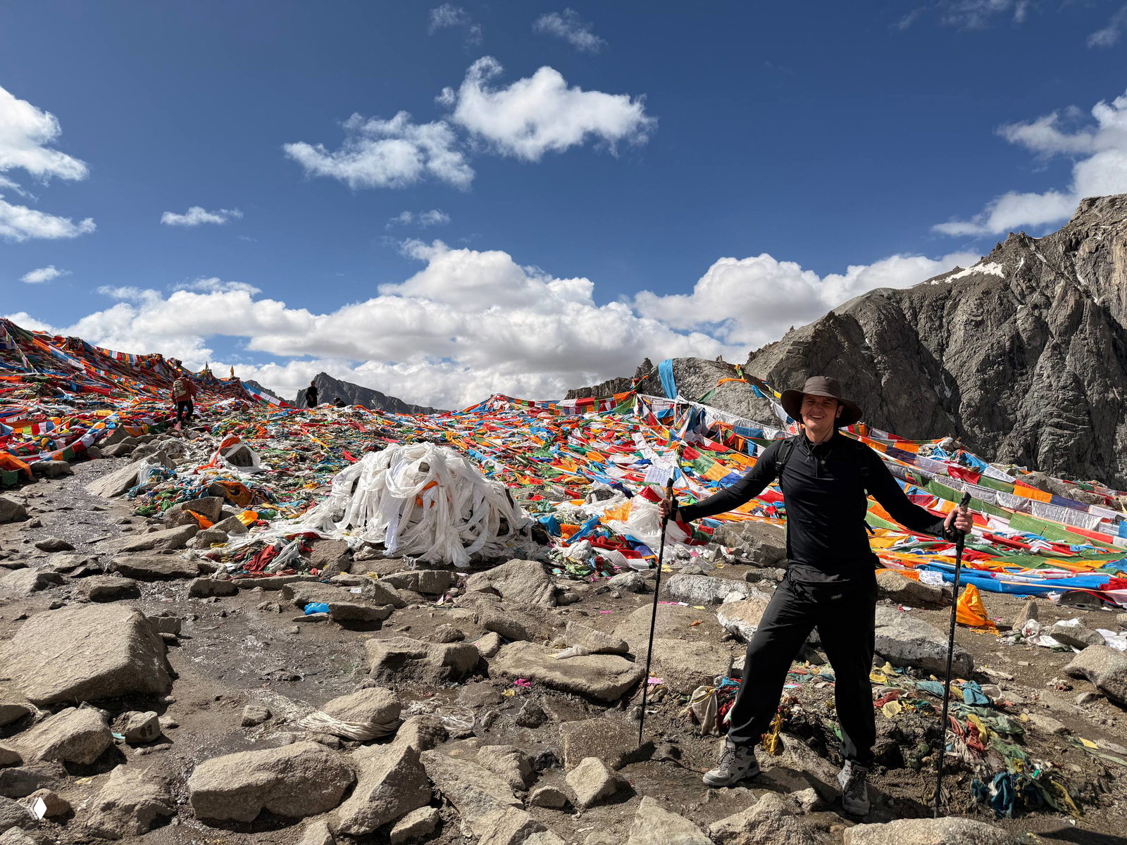 Автор стоит на каменистом склоне у большого скопления разноцветных молитвенных лент и флажков, горы и облачное небо за спиной.