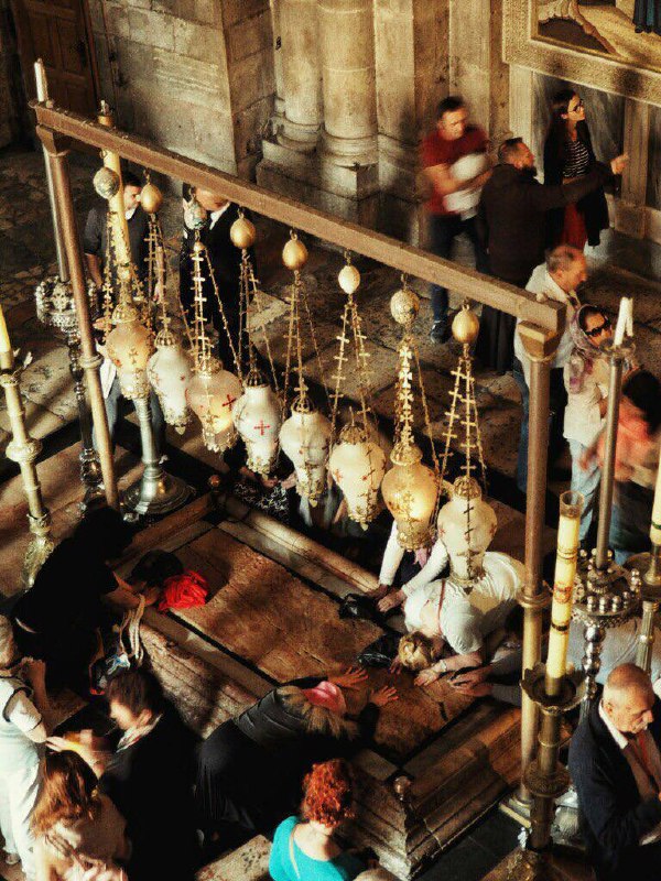 Паломники у мраморной плиты внутри храма: люди склоняются и целуют святыню, вид сверху на ритуальную зону и лампы над ней.