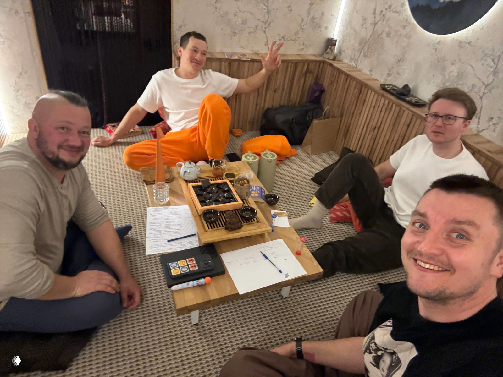 Четверо мужчин сидят на полу вокруг низкого столика с чайной посудой и заметками — мини-группа обсуждения и совместной практики.