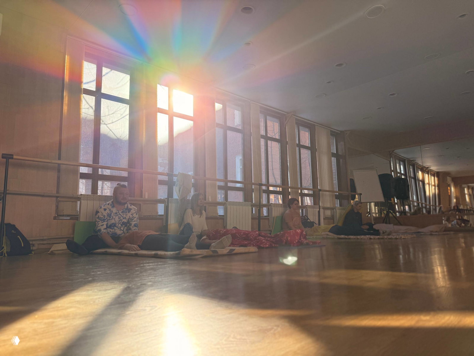 Большой зал с людьми, лежащими и сидящими на матах у окон, солнечные лучи проходят сквозь стекло и создают радужные блики.