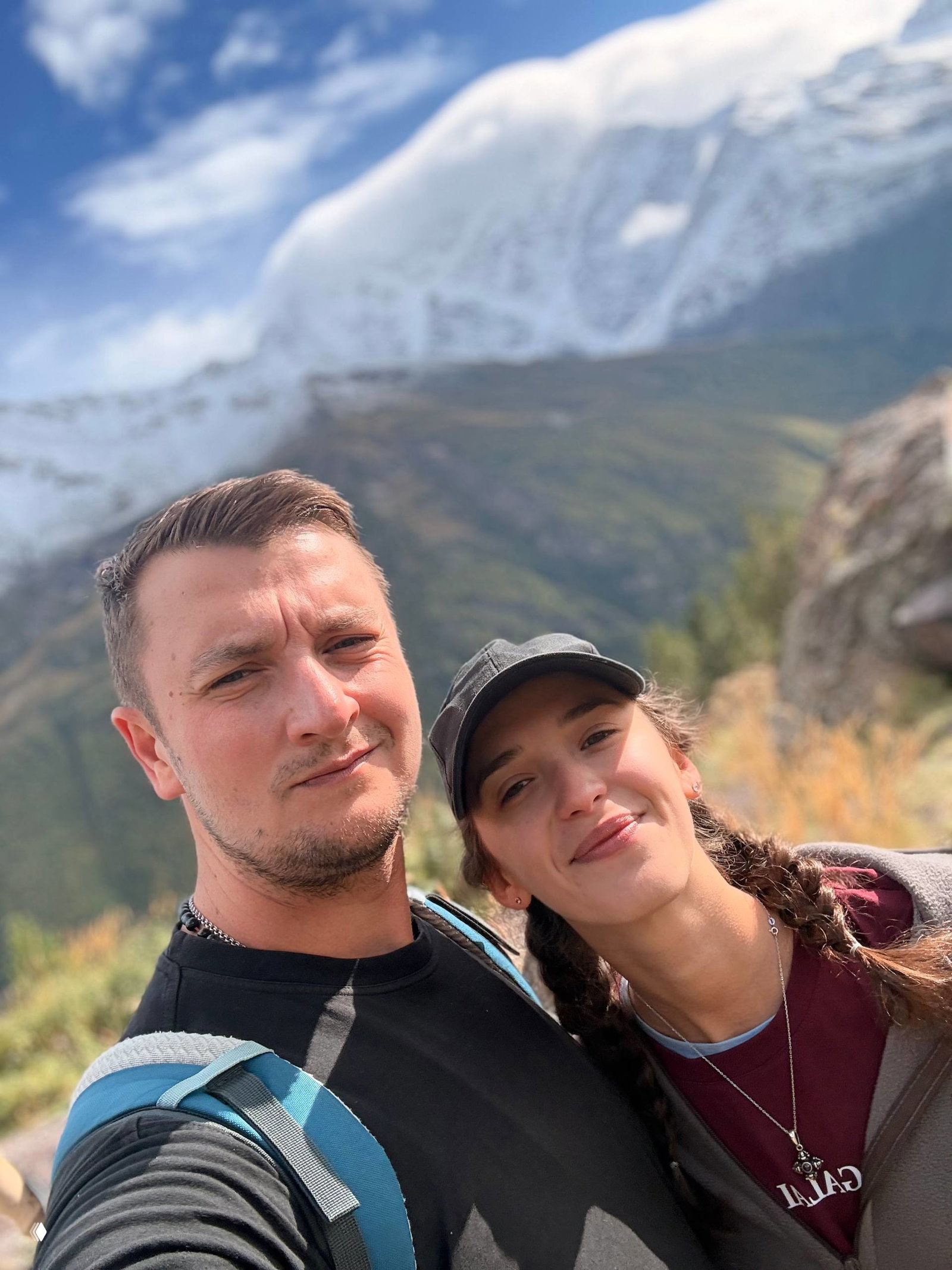 Селфи пары на фоне гор: мужчина и женщина в повседневной экипировке, горный пейзаж и ясное небо создают атмосферу путешествия и близости.