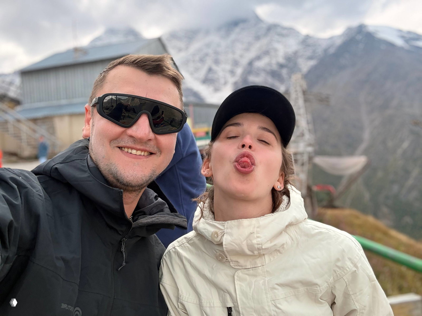 Селфи на горнолыжной станции: мужчина в тёмной куртке улыбается, женщина в кепке шутит и показывает язык, на заднем плане заснеженные склоны.