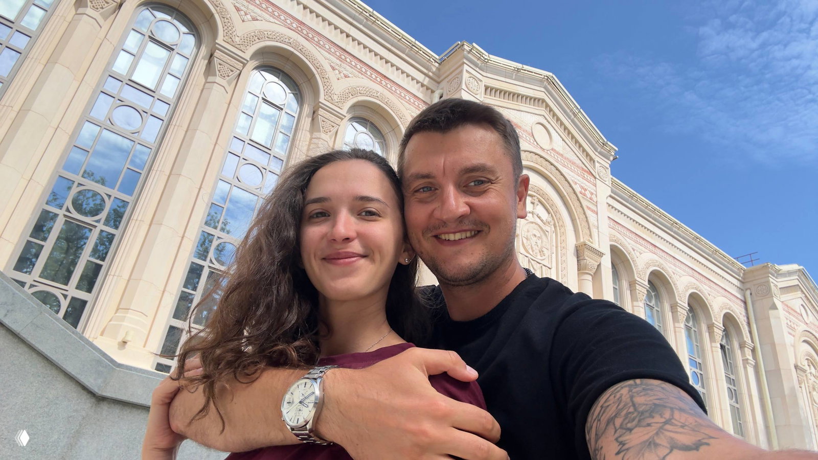 Пара на фоне архитектурного фасада: мужчина обнимает женщину рядом с большим зданием с арочными окнами, дневной свет подчёркивает лица.