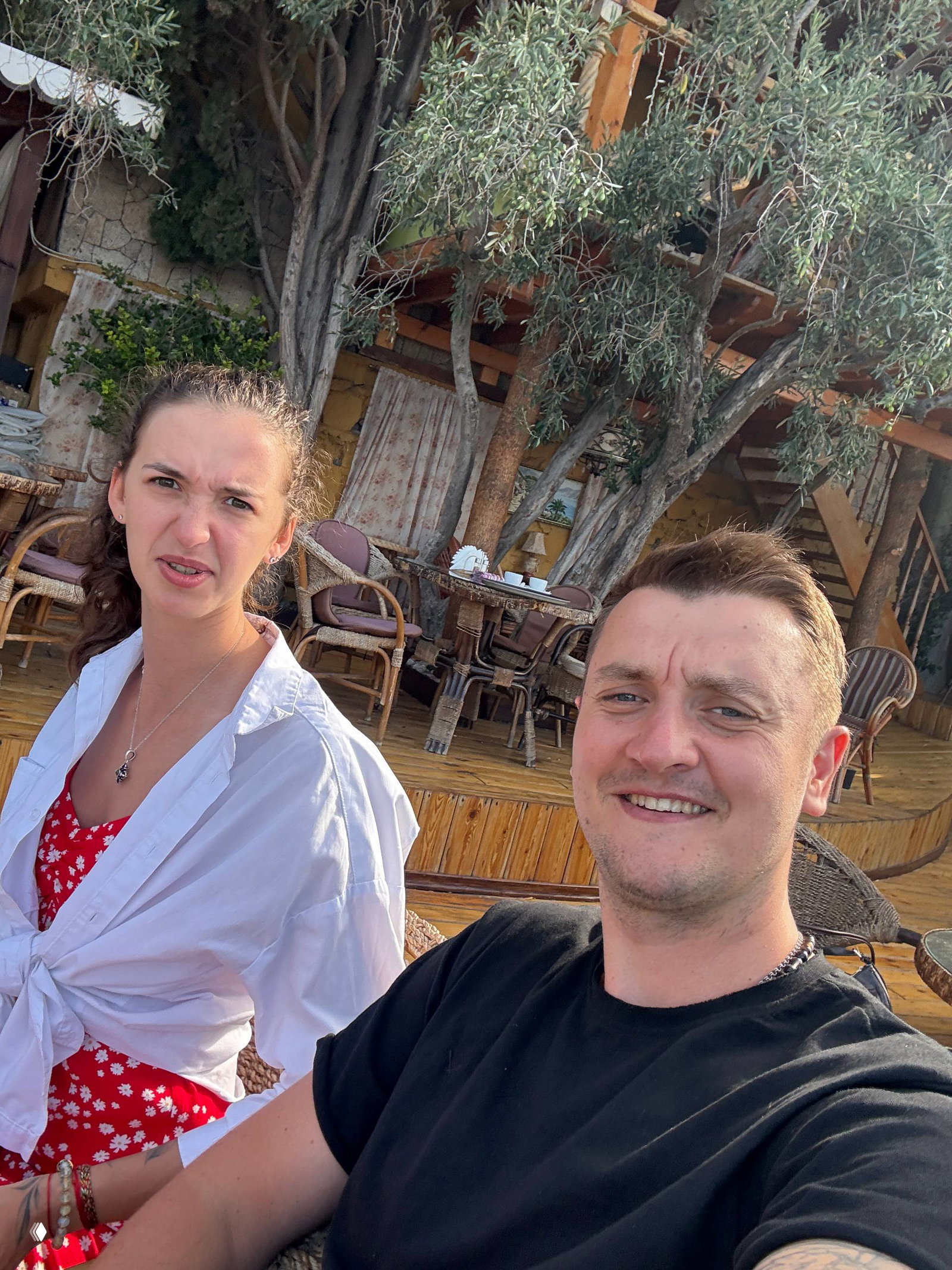 Пара на открытой террасе: женщина в ярком платье и мужчина улыбаются за столиком, вокруг деревянная мебель и зелень, повседневная сцена.